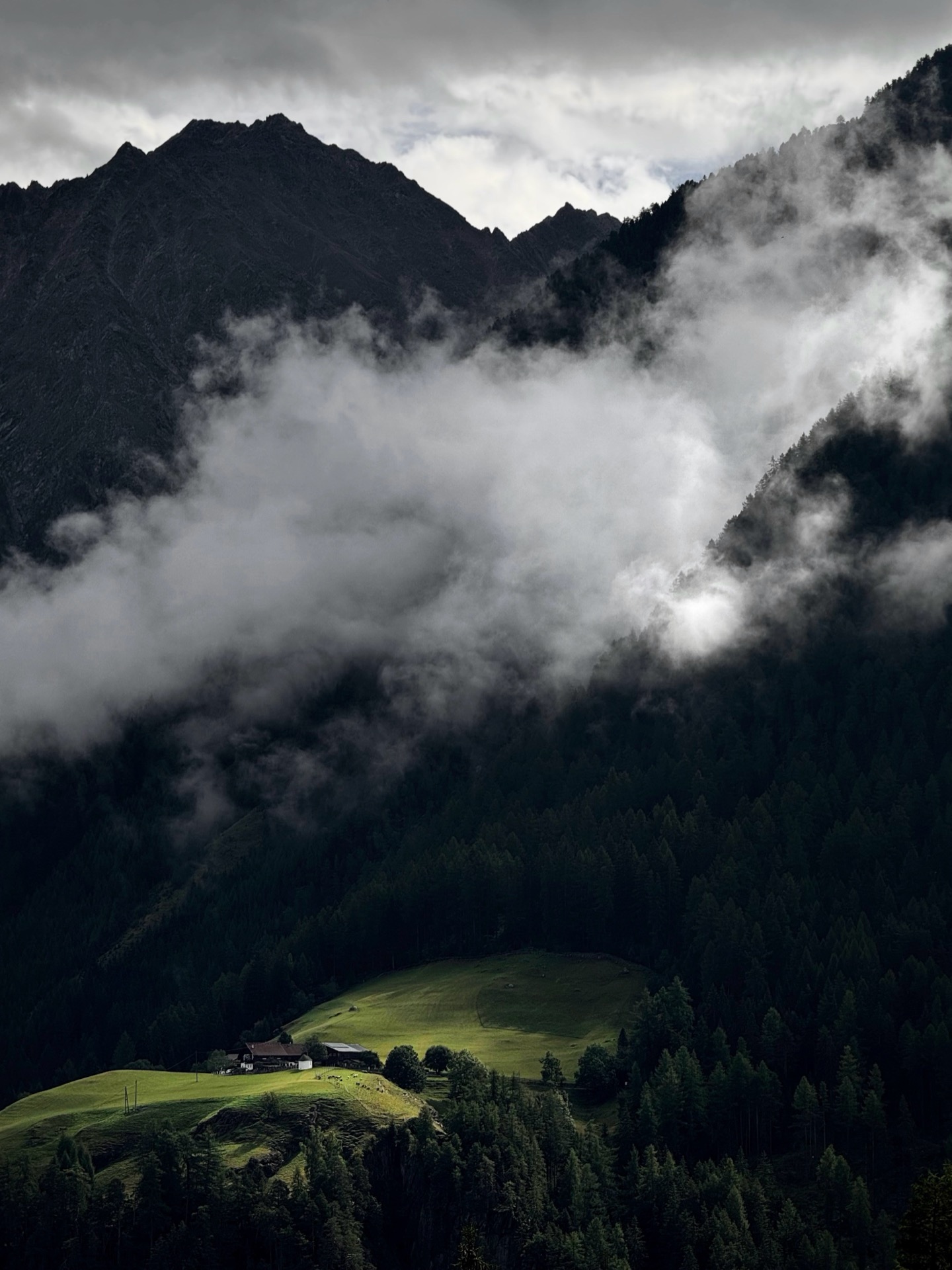 A lone farmhouse on a green shoulder, cloud pouring through the conifer slopes above it